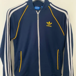 Adidas marinblå vindjacka med gula detaljer - Snygg marinblå tracktop från Adidas med klassiska vita  och gula detaljer. Riktigt bra som att gå på fotboll med, eller i vardagen. Några slitningar men inget som märks. Står S men passar som XS