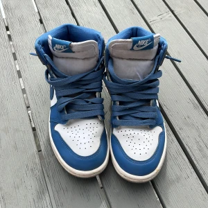 Nike Air Jordan 1 High blå/vit/grå - Säljer ett par Nike Air Jordan 1 High med blå och vita paneler samt grå mockadetaljer. Klassisk high-top siluett med snörning, perforerad tå och ikoniska Air Jordan-loggan på sidan. Skorna har platt sula och är tillverkade i skinn och mocka. Strl 34