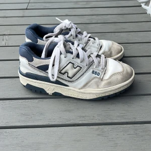 New Balance 550 vita och marinblå sneakers strl 36 (junior) - Säljer ett par New Balance 550 sneakers i vitt och marinblått med klassisk retrodesign. Skorna har snörning, rund tå och en mix av skinn och mocka i ovandelen. Snygga detaljer med NB-logga på hälen och sidan samt robust sula för extra komfort. Junior strl 36.