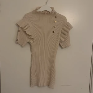 Beige ribbad blus med volang från raglady  - Superfin beige ribbad bluS med volangdetaljer på ärmarna och snygga guldknappar längs axeln. Hög hals med ruffle-kant och korta ärmar. Perfekt för dig som gillar en söt men stilren look. Storlek S/XS