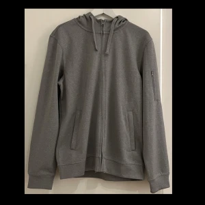 Grå hoodie med dragkedja från Clique - Snygg grå hoodie från Clique i storlek M. Tröjan har hel dragkedja, huva med snörning och två fickor fram samt en extra ficka med dragkedja på ärmen. Perfekt basic-plagg med skön passform och stilren look.