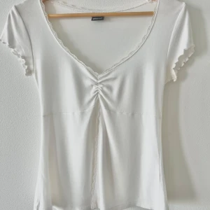 Vit v-ringad topp från Gina Tricot - Supersöt vit topp från Gina Tricot med v-ringning och spetskant. Toppen har korta ärmar med vågig kant, rynkdetalj framtill och en feminin passform. Perfekt för dig som gillar en enkel men ändå romantisk stil.