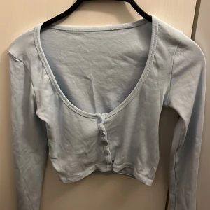 Ljusblå croppad topp med knappar Missay - Supersöt ljusblå croppad topp från Missay med lång ärm och djup rundringning. Toppen har fyra knappar framtill som snygg detalj och är gjord i mjuk bomull med lite stretch för en skön passform.