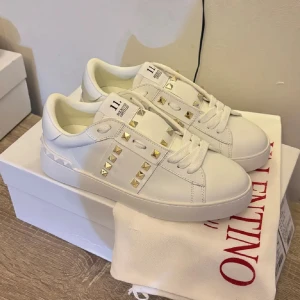 Valentino Rockstud vita sneakers(Nya) - Snygga vita Valentino Garavani Rockstud sneakers i skinn med ikoniska guldiga nitar och coola detaljer på sidorna. Klassisk rund tå, platt sula och breda vita snören. Perfekta för dig som vill sticka ut med exklusiv streetstyle. allt finns kvar!❤️