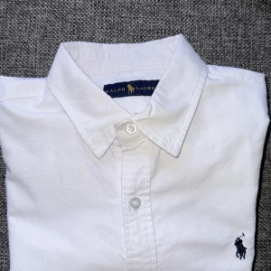 Vit skjorta från Ralph Lauren - Klassisk vit skjorta från Ralph Lauren med broderad marinblå logga på bröstet. Skjortan har lång ärm, knappar framtill och en snygg krage. Tillverkad i bomull för en fräsch och stilren look. Skjortan är i stort sett helt oanvänd***. Säljer den pga att den var för liten och att jag råkade vänta för länge med att skicka tillbaka den.