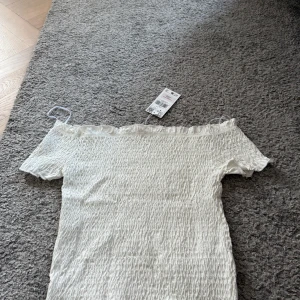 Vit offshoulder topp från Mango - Säljer en vit topp från Mango i storlek S. Toppen  har offshoulder-design med smockad struktur och tunna axelband. Materialet är mjukt och stretchigt, perfekt för sommarens varma dagar. Söt volang upptill