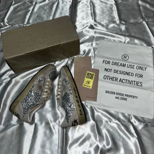 Silverglittriga Golden Goose Ball Star - Säljer ett par Golden Goose Ball Star sneakers med silverglittriga paneler, beige mockadetaljer och vit stjärna på sidan. Skorna har snörning, rund tå och platt sula. Märkets logga syns tydligt på hälen och innersulan. Perfekt för dig som gillar att sticka ut.