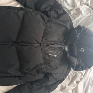 Svart pufferjacka från Sail Racing - Säljer en svart pufferjacka från Sail Racing med huva och dragkedja framtill. Jackan har en clean look med diskret logga på bröstet och praktiska fickor med dragkedja. Perfekt för kalla dagar när du vill hålla värmen och ändå se snygg ut.