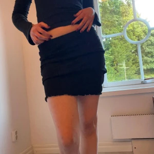 Svart tight kort kjol med volang - Säljer en svart kort kjol med tight passform. Kjolen har vågiga sömmar längs framsidan och en volangkant nedtill som ger en cool look. Perfekt för dig som gillar minimalistisk stil med en twist.