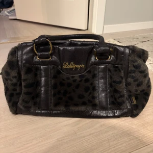Leopardmönstrad handväska från Lollipops - Cool handväska från Lollipops i svart skinnimitation med mjuka pälsdetaljer och leopardmönster. Väskan har guldiga metalldetaljer, dubbla handtag och ett ljust innerfoder med tryck. Perfekt för dig som vill sticka ut med en unik accessoar.
