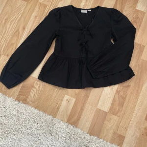 Svart blus med knyt och v-ringning - Svart blus med v-ringning och knyt framtill. Långa puffiga ärmar och lätt utställd nertill för en snygg siluett. Perfekt att styla med jeans eller kjol för en trendig look. Aldrig använd❤️