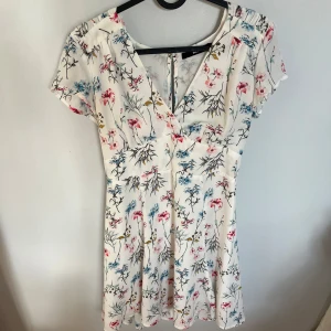 Vit blommig kortklänning Bik Bok - Supersöt vit kortklänning från BikBok med blommigt mönster i rosa, blått och gult. Klänningen har v-ringning, korta ärmar och markerad midja. Använd fåtal gånger men inga tecken på användning och inga defekter. Perfekt nu till sensommaren eller hösten med en stickad tröja över. 