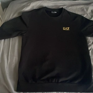 Svart EA7 Emporio Armani sweatshirts Armani tröja storlek M - Svart sweatshirt från EA7 Emporio Armani med gul logga på bröstet och diskreta guldfärgade detaljer på ryggen. Klassisk rund hals och långa ärmar. Perfekt för dig som gillar stilrena och exklusiva plagg med en sportig touch.