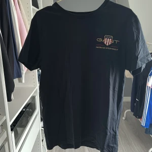 Svart GANT t-shirt med broderad logga - Svart t-shirt från GANT i bomull med broderad logga. Klassisk rund halsringning och korta ärmar. Snygg och stilren modell som passar perfekt till jeans eller shorts.