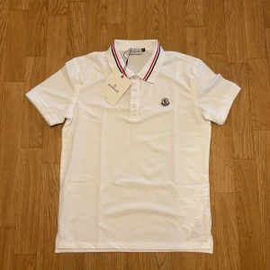 Vit Moncler pikétröja - Vit pikétröja från Moncler med klassisk krage och knappar framtill. Kragen har snygga röda och svarta ränder och Moncler-logga är broderad på bröstet. Tröjan är kortärmad och tillverkad i bomull för en fräsch och stilren look.