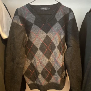 Rutig svart och grå tröja Clique - Snygg tröja från Clique i svart och grått med klassiskt rutig mönster och röda detaljer. V-ringad hals och långa ärmar. Perfekt för dig som gillar en stilren men ändå lite preppy look.