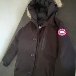 Svart dunjacka från Canada Goose - Säljer en svart dunjacka från Canada Goose med klassisk patch på ärmen och stor huva med fuskpäls. Jackan har dragkedja och knappar framtill, samt flera fickor. Perfekt för kalla vinterdagar och riktigt snygg streetstil.