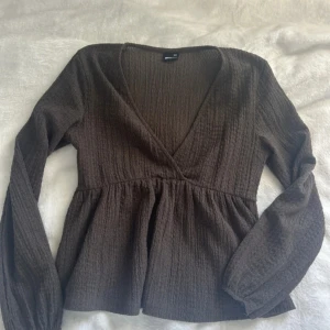 Brun omlottopp med v-ringning - Supersnygg brun omlottopp med v-ringning och långa ärmar. Toppen har en lätt rynkad struktur och markerad midja som ger en fin siluett. Perfekt att styla med jeans eller kjol för en trendig look.