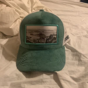 Grön och vit keps från Pianotti - Snygg truckerkeps från Pianotti med grön front och skärm i mjukt tyg, vit mesh baktill och broderad logga på sidan. Framsidan har en cool svartvit bild med turkos ram. Perfekt för dig som vill sticka ut med en unik accessoar.