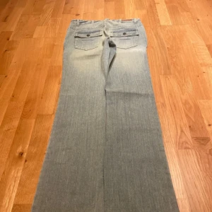 bootcut jeans - Säljer ett par ljusblå jeans med bootcut passform och snygga fickor med lock och knappar bak. Jeansen har slitningar framtill och klassisk femficksdesign. Perfekta för dig som gillar en retro vibe och vill ha ett par jeans som sticker ut. färgen är blå/grå