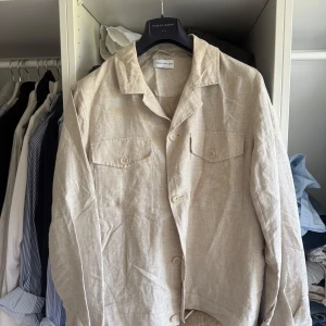 Beige overshirt från These Glory Days - Snygg beige overshirt från These Glory Days i linne. Jackan har klassisk krage, två bröstfickor med lock och knappar framtill. 
