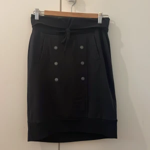 Svart kjol med knappar House of Lola XS - Svart kortkjol från House of Lola med snygga dekorativa knappar framtill och knyt i midjan. Kjolen har fickor på sidorna och en bredare kant nedtill för en cool siluett. Perfekt för dig som gillar stilrena och enkla plagg med en twist. 94% bomull och 6% elastan. Storlek XS men passar även S