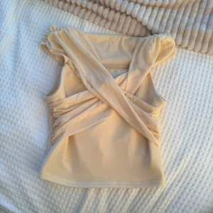 Beige omlott ärmlös topp - Supersnygg beige topp med omlott-detalj framtill och breda axelband. Ärmlös modell i mjukt, stretchigt material som sitter skönt och ger en cool look. Perfekt att styla med jeans eller kjol för en trendig vibe.