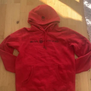 Röd hoodie från Sail Racing - Röd hoodie från Sail Racing med svart logga på bröstet. Tröjan har huva med snörning och en stor magficka framtill. Tillverkad i mjuk bomull, perfekt för dig som gillar en avslappnad och sportig stil. Knappt använd och passar M-S. (Pris går att diskutera)