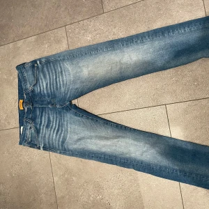 Blå jeans från Jack & Jones - Säljer ett par klassiska blå jeans från Jack & Jones med snyggt slitna detaljer och femficksmodell. Jeansen har normal passform och raka ben, tillverkade i bomull med en skön jeanskänsla. Perfekta för dig som gillar en tidlös och avslappnad stil.