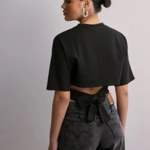 Svart knyt topp nelly - Svart croppad topp med korta ärmar och rund halsringning. Baksidan har en öppen detalj med breda band som knyts i en rosett i midjan. Enkel och stilren design som ger en cool twist till outfiten.
