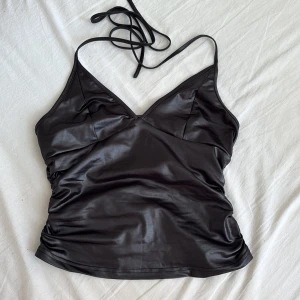 Svart faux skinn linnetopp från Cider - Säljer en svart linnetopp från Cider i glansigt faux skinn-material. Toppen har smala axelband som knyts i nacken, öppen rygg, v-ringning och rynkade sidor för en snygg passform. Perfekt för dig som vill sticka ut med en edgy look.