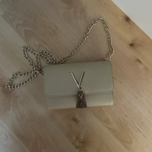 Beige axelväska från Valentino - Snygg beige axelväska från Valentino med guldfärgad kedja och V-detalj framtill. Väskan har en stilren design med tofsdetalj och är tillverkad i skinnliknande material. Perfekt för dig som vill ha en trendig väska