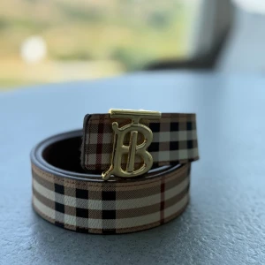 Burberry bälte - Snyggt bälte från Burberry med klassiskt rutigt mönster i beige, svart, vitt och rött. Bältet har ett stilrent guldigt spänne med Burberrys ikoniska monogram. Perfekt accessoar för att lyfta din outfit med en lyxig touch. 