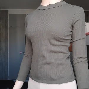 Ribbad grå polotröja från Primark - En ribbad grå polotröja från Primark med lång ärm och hög hals. Tröjan är i stretchigt material och har en enkel, clean look som funkar till många outfits. Perfekt för lager-på-lager under kyliga dagar.