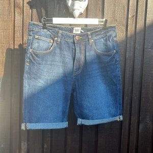 Blå jeansshorts från 157 - Klassiska blå jeansshorts från 157 med uppvikta benslut och femficksmodell. Snygga kontrastsömmar och läderpatch bak i midjan. Perfekta för varma dagar och enkel att matcha med t-shirt eller hoodie.