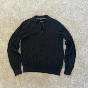 Mörkgrå stickad tröja Riley half zip - Mörkgrå stickad tröja från Riley i bomull med snygg half zip-dragkedja i guldton. Tröjan har ribbade muddar och krage, samt lång ärm. Perfekt för dig som gillar en clean och stilren look. Storlek M men passar S