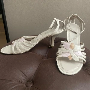 Beige sandaler med klack och blomdetalj - Snygga sandaler från Rocco Barocco i beige skinn med smala remmar och hög klack. Framtill sitter en stor blomma i beige och ljusrosa nyanser som ger en söt och unik touch. Perfekta för dig som vill sticka ut med en feminin och trendig stil. OBS! Klisterlappen togs bort från sulan och en liten del av ytan följde med (se bild). Inget som syns när skon används och påverkar inte funktionen.