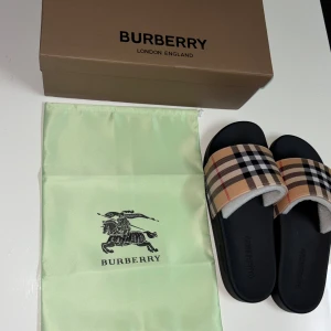 Burberry sandaler - Svarta sandaler från Burberry med bred rem i det ikoniska beige, svarta och röda rutmönstret. Snygg och stilren design med platt sula och rund tå. Tillverkade i Italien och perfekta för en trendig look.
