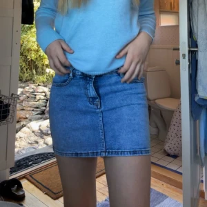 Blå jeanskjol i klassisk modell - Säljer en snygg blå jeanskjol. Kjolen är kort och har en enkel design med knapp och dragkedja framtill. Perfekt till sommaren och lätt att matcha med olika toppar. Endast testad 