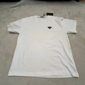 Vit t-shirt från Prada med logga - Vit t-shirt från Prada i klassisk modell med korta ärmar och rund hals. På bröstet sitter en ikonisk svart och silverfärgad Prada-logga i metall. T-shirten är gjord i mjuk bomull och har en clean, stilren look som passar perfekt till jeans eller shorts.