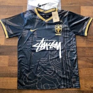 Svart Brasilien x Stüssy x Nike tröja - Snygg svart fotbollströja från Nike i samarbete med Stüssy och Brasilien. Tröjan har guldiga detaljer vid krage och ärmslut, samt ett unikt grafiskt mönster. Stüssy-loggan är tryckt i vitt över bröstet och Brasilien-emblemet i guld.