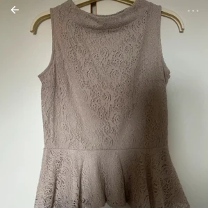 Beige spetsblus från Lindex - Supersöt beige blus från Lindex i storlek S. Toppen är ärmlös och har ett peplumliv med volang nertill. Hela toppen är täckt av vacker spets med blommönster och har en mjuk, feminin look. Perfekt att styla med jeans eller kjol för en chic vibe.