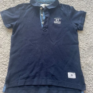 Marinblå pikétröja Hampton Republic - Snygg marinblå pikétröja från Hampton Republic med klassisk krage, tre knappar och broderad logga på bröstet. Tröjan har korta ärmar och detaljer i ljusblått tyg vid knäppningen. Perfekt för en stilren och sportig look.