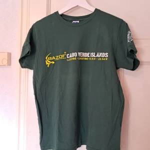 Grön t-shirt Cabo Verde Islands Russell - Snygg mörkgrön t-shirt från Russell med tryck 'Cabo Verde Islands' och koordinater i vitt och gult framtill. Stor tribal-inspirerad sköldpadda i gult på ryggen och mindre tryck på ärmen. Klassisk passform och rund hals, tillverkad i mjuk bomull. Köpt i Kap Verde.