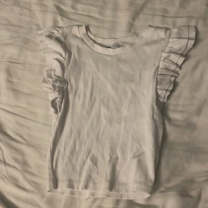 Vit ribbad topp med volangärm från Zara - Supersöt vit ribbad topp från Zara med volangdetaljer på axlarna. Toppen är ärmlös och har en rund halsringning. Perfekt för dig som gillar en feminin och trendig stil. Materialet är mjukt och stretchigt för en bekväm passform. Är kort i modellen. 