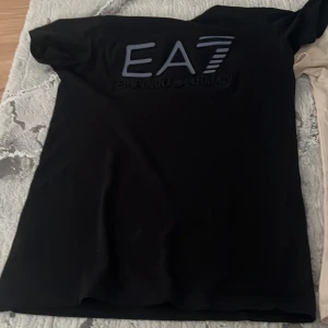Svart EA7 Emporio Armani t-shirt - Svart t-shirt från EA7 Emporio Armani med stor, upphöjd logga på bröstet. Klassisk passform och korta ärmar, perfekt för dig som gillar stilrena och exklusiva plagg. Materialet känns mjukt och följsamt mot huden.