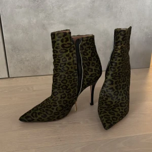 Leopardmönstrade stövletter med klack - Stövletter med leopardmönster i grönt och svart. Skorna har spetsig tå, hög smal klack och dragkedja på sidan med guldfärgad detalj. Materialet är pälsliknande. Inte för den fega. 