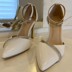 Vita klackar med strass  - Snygga vita klackar  med spetsig tå, hög klack och elegant struktur i materialet. Skorna har en smal rem med glittrande strassdetaljer och justerbar ankelrem. De har använts 1 gång bara och ser helt nya ut. 