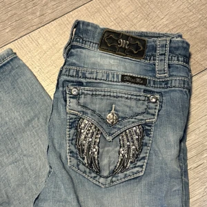 Miss Me ljusblå jeans med bling - Fick dom av min kusin och vill tjäna lite pengar Snygga ljusblå jeans från Miss Me med blingade detaljer och broderade vingar på bakfickan. Jeansen har raka ben, dekorativa knappar och coola sömmar. Perfekta för dig som vill sticka ut med lite extra detaljer.