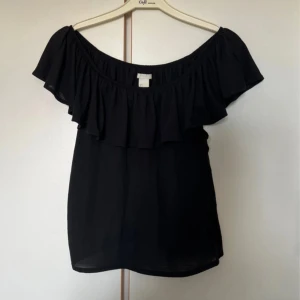 Svart offshoulder volangtopp H&M - Svart offshoulder topp från H&M med bred volang runt hela ringningen. Tunn och luftig modell som sitter snyggt över axlarna. Perfekt för dig som gillar en enkel men trendig look.